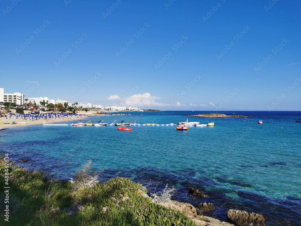 Protaras. Famagusta area. Cyprus. The coast of the Mediterranean Sea with green plants, a view of the sandy beach of Fig Tree Bay and the island in the background of a blue sky with clouds.