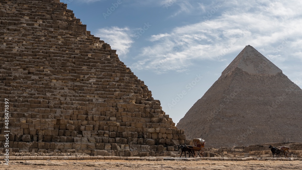 The two great pyramids of Giza- Cheops and Chephren against the blue sky. The ancient masonry walls are visible. At the foot are horses harnessed to carts. Egypt The two great pyramids of Giza- Cheops and Chephren against the blue sky. The ancient masonry walls are visible. At the foot are horses harnessed to carts. Egypt