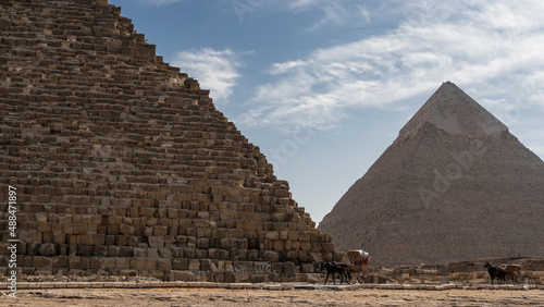The two great pyramids of Giza- Cheops and Chephren against the blue sky. The ancient masonry walls are visible. At the foot are horses harnessed to carts. Egypt