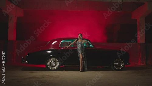 Beautiful Elegant Young Woman Standing , Dancing and Flirting near old retro classic car. Old fashioned luxury style concept . Rich fashion lifestyle . Luxury dress with fabric glitter, spangles . photo