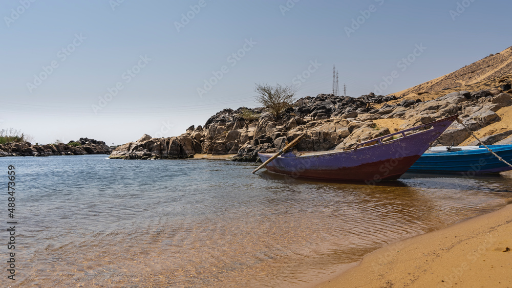 Old painted wooden boats are moored at the sandy shore of the Nile. Ripples on clear water. Picturesque boulders on the shore. Blue sky. Egypt