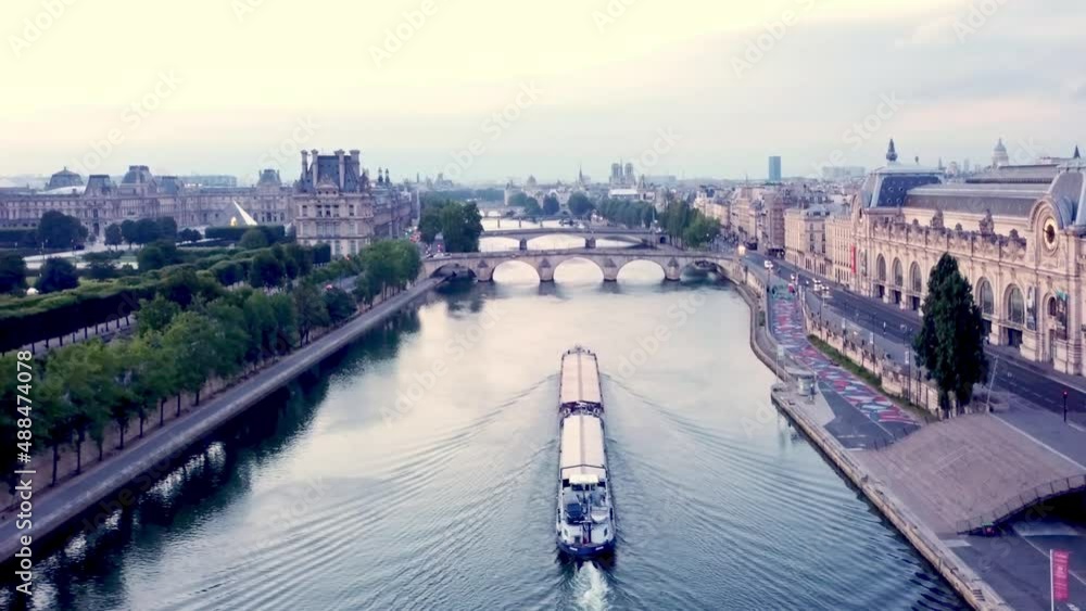 Aerial moving (back up) over Seine river, with a barge moving opposite. View on Louvre Museum, Notre-Dame in Background, Orsay. Paris, France