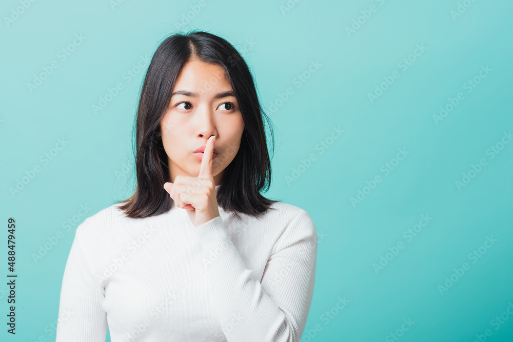 Young beautiful Asian woman holding index finger on her mouth lips, Portrait female hush silence, studio shot isolated on a blue background, Gesture of shhh Young beautiful Asian woman holding index finger on her mouth lips, Portrait female hush silence, studio shot isolated on a blue background, Gesture of shhh