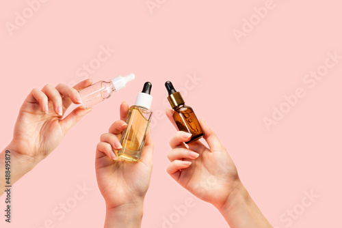 Three female's hands hold glass multi-colored bottles with oil and serum. Pink background, close-up. Copy space. The concept of care cosmetics for any skin type photo