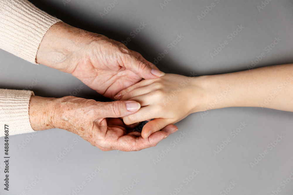 Fototapeta premium Grandmother is holding her granddaughter's hand. Hands close-up. Flat lay. Gray background. The concept of caring for pensioners