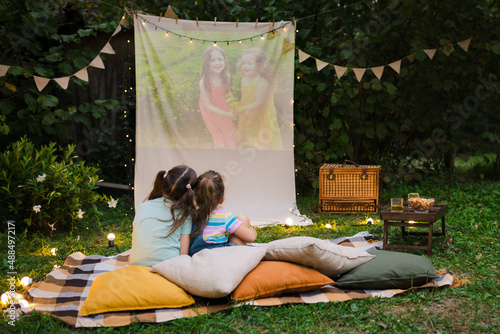 Open air cinema. Backyard Family outdoor movie night with kids. Sisters spending time together and watching cimema at backyard. DIY Screen with film. Summer outdoor weekend activities with children