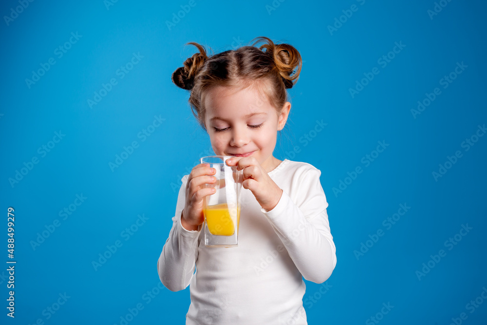 little girl with a funny hairstyle in a white T-shirt holds a glass with juice in her hands. blue background. useful products for children. space for text. High quality photo little girl with a funny hairstyle in a white T-shirt holds a glass with juice in her hands. blue background. useful products for children. space for text. High quality photo