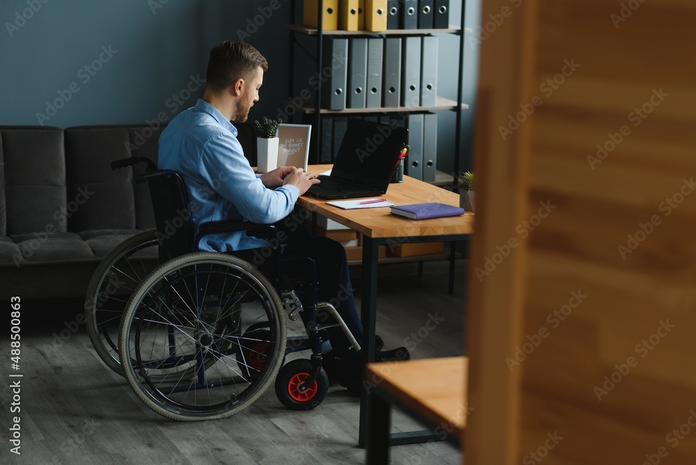 Young man with special needs in casual clothes working on wireless laptop. Male freelancer working from home while sitting in wheelchair. Young man with special needs in casual clothes working on wireless laptop. Male freelancer working from home while sitting in wheelchair.