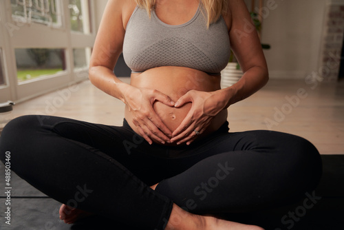 Pregnant Caucasian female sitting cross legged on yoga mat while creating a heart with her hands on her belly. Healthy lifestye in modern home. photo