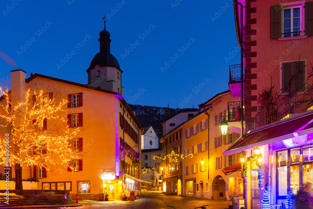 Festive atmosphere of narrow streets of small Alpine mountainous town of Brig during winter Christmas season with bright lighting and traditional decorations at night, Switzerland
