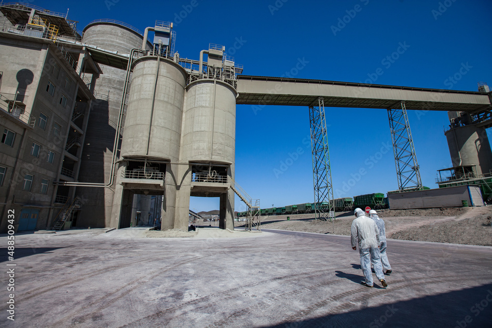 Obraz premium Modern cement plant in desert. Two workers in protective overalls or work wear and cement silo on deep blue sky. Mynaral, Kazakhstan. 
