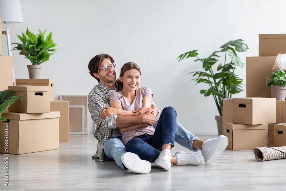 Glad millennial caucasian male in glasses and female in casual hugs, sits on floor among cardboard boxes Glad millennial caucasian male in glasses and female in casual hugs, sits on floor among cardboard boxes