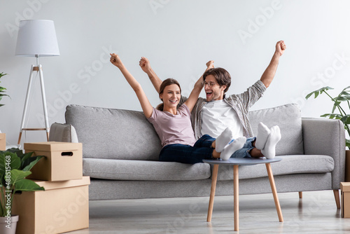 Glad happy young caucasian husband and wife in casual raise their hands up celebrating moving on couch