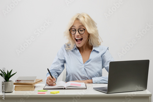 Surprised businesswoman working on laptop and taking notes in notebook, surprisingly reading her notations at workplace
