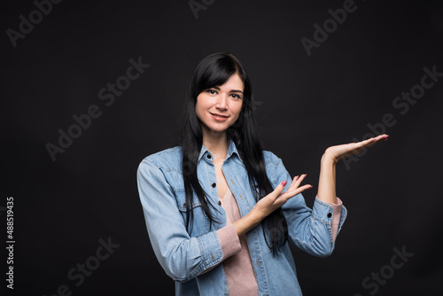 Attractive caucasian brunette girl in a shirt pointing with her hand to the palm with copy space for advertising isolated on black studio background.