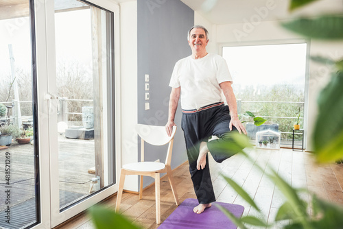 Smiling senior man standing on one leg by chair at home