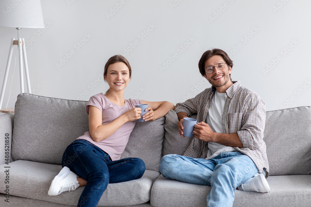 Happy glad millennial caucasian guy in glasses and woman in casual rest on sofa, drinking coffee Happy glad millennial caucasian guy in glasses and woman in casual rest on sofa, drinking coffee