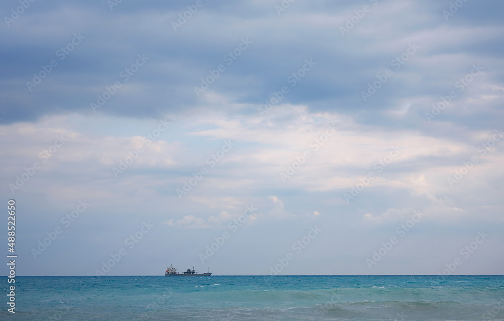 Cargo ship in calm blue Mediterranean in Anatalya. Traveling along the Mediterranean Sea, view of rocky coast of Antalya. In distance, cargo ships are visible.