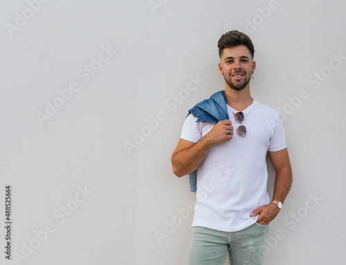 A nice style young man smiling looking at the camera while leaning in a white wall and have a jacket in his right shoulder.