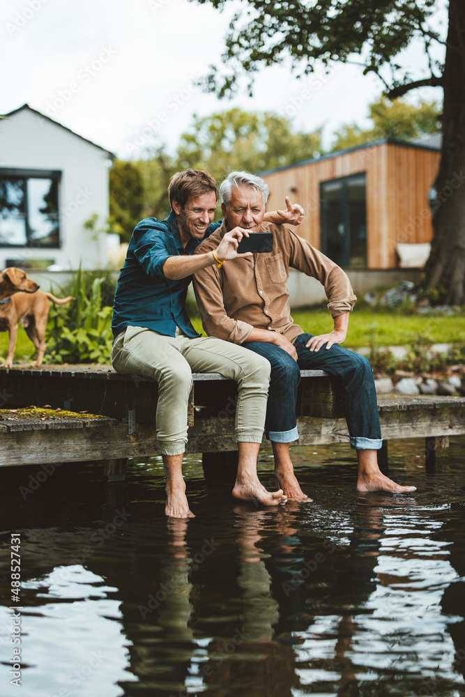 Smiling man taking selfie with father through smart phone on jetty by lake Smiling man taking selfie with father through smart phone on jetty by lake