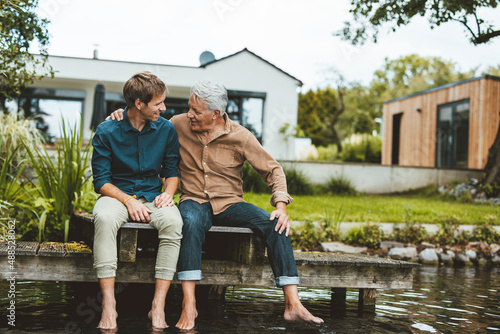 Senior man talking with son sitting on jetty by lake at backyard