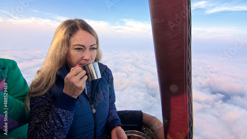 Woman eat sandwich and drinks tea and coffee in flight. Adventure on hot air balloon. Burner directing flame into envelope. Fly in morning blue sky above cloud. Girl stand in basket hot air balloon. photo