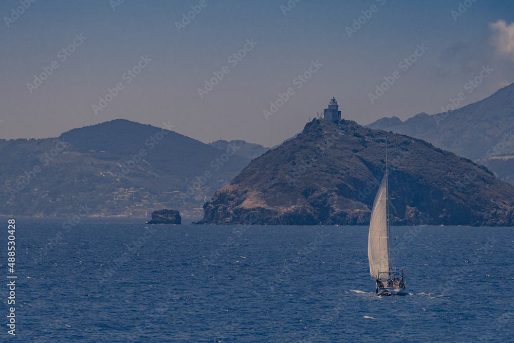 Sea scene with a sailboat heading for land. An old lighthouse built on top of a small rocky island overlooks the subject. The sky is clear and there is a light mist in the air, suggesting a hot, muggy Sea scene with a sailboat heading for land. An old lighthouse built on top of a small rocky island overlooks the subject. The sky is clear and there is a light mist in the air, suggesting a hot, muggy