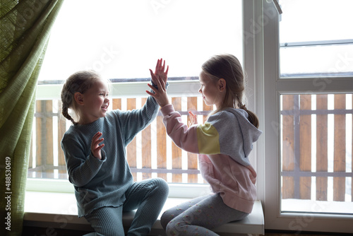 two girls are playing patty-cake, sitting on the windowsill near the big window photo