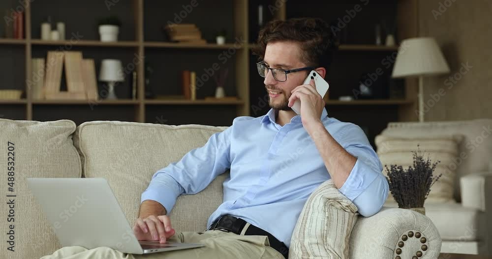 Busy young male freelancer wear glasses sit on sofa engaged in multitask job using gadgets. Millennial man work from home with comfort hold business conversation talk on phone type on laptop keyboard Busy young male freelancer wear glasses sit on sofa engaged in multitask job using gadgets. Millennial man work from home with comfort hold business conversation talk on phone type on laptop keyboard