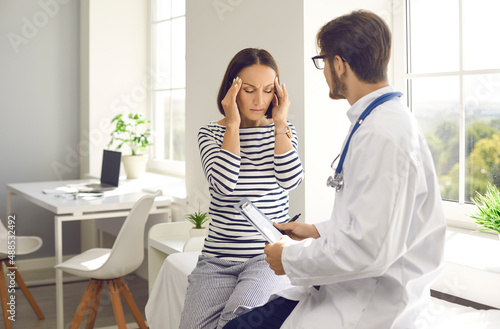 Tired senior woman complains to doctor of severe headaches during visit or consultation at clinic. Young male doctor records patient's symptoms during medical examination in hospital room.