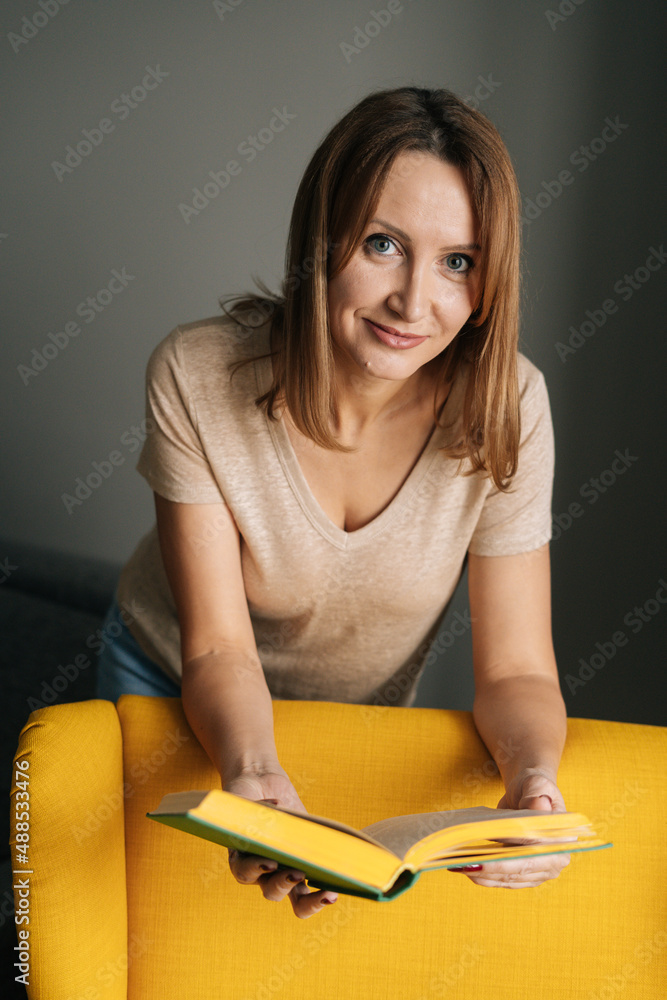 Vertical shot of confident blonde woman standing near yellow chair reading paper book in dark room with modern interior, smiling looking at camera, on background of bookshelves. Vertical shot of confident blonde woman standing near yellow chair reading paper book in dark room with modern interior, smiling looking at camera, on background of bookshelves.