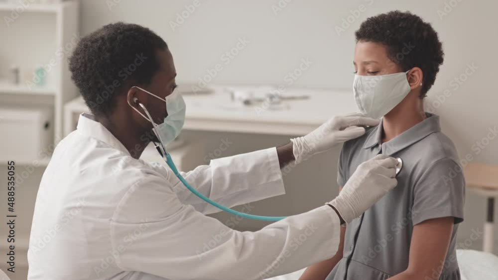 Waist-up slowmo shot of African-American doctor listening heart of African-American schoolgirl with stethoscope both wearing face masks Waist-up slowmo shot of African-American doctor listening heart of African-American schoolgirl with stethoscope both wearing face masks