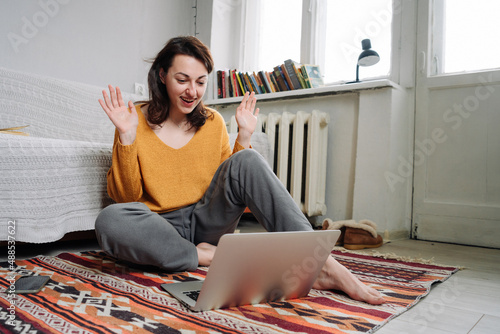 A young girl sitting on the floor looks at her laptop, smiles and waves with both hands photo