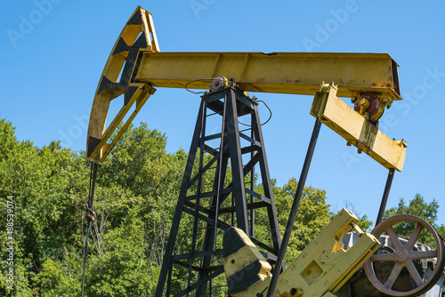 Close-up of a yellow old oil rig pumping oil from a well against a background of green trees and blue sky. View from below.