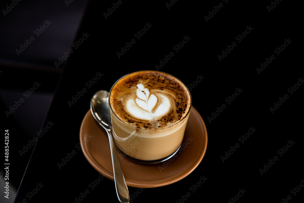 Glass of cappuccino with latte art on saucer and with spoon on the black table