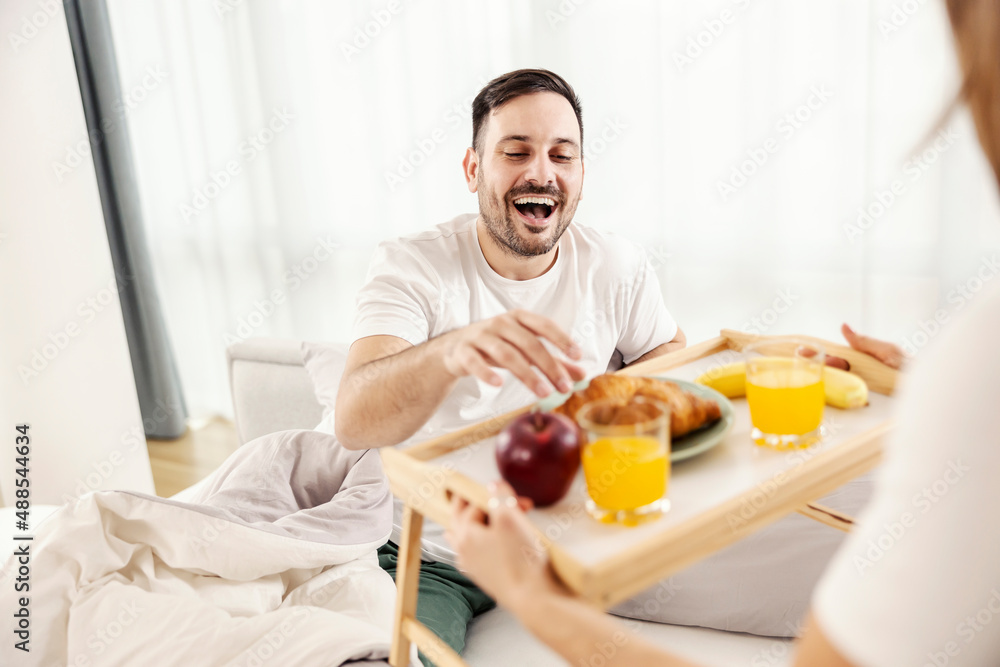 A happy man having breakfast in the bed brought by his wife at their cozy home. A happy man having breakfast in the bed brought by his wife at their cozy home.