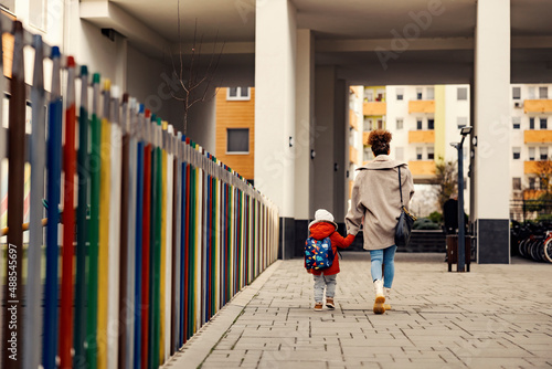 A nanny holding hands with little boy with a schoolbag and taking him to kindergarten.