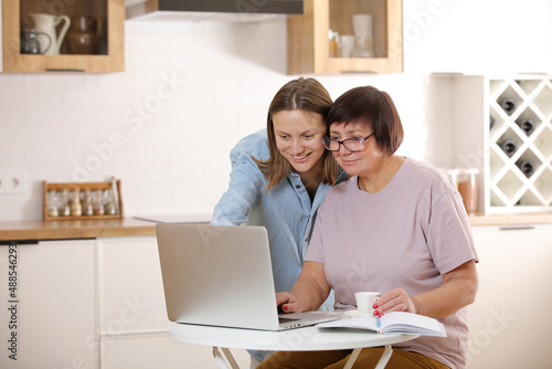 young woman teaching senior mother to use internet on laptop at home. daughter helps her elderly mother figure it out online with her personal account, teaches at modern gadget indoors.