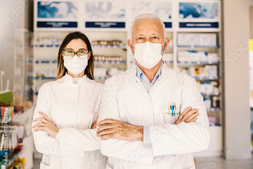 Young and old pharmacy workers with mask posing in pharmacy.