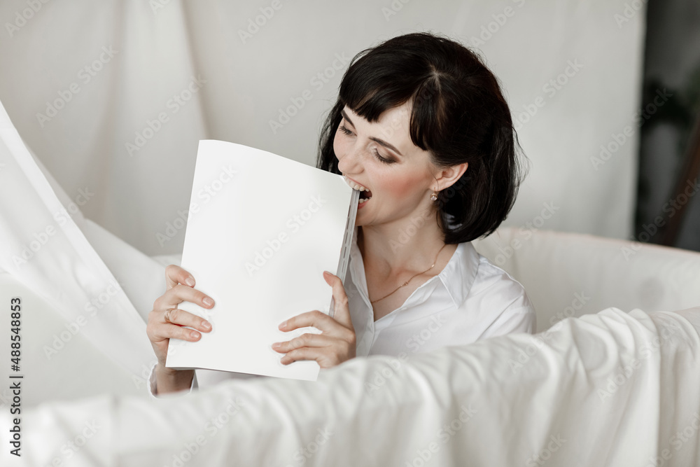 brunette businesswoman bites books moke up, reads or keeps. girl with dark short hair in a white shirt, studio isolated portrait emotions brunette businesswoman bites books moke up, reads or keeps. girl with dark short hair in a white shirt, studio isolated portrait emotions