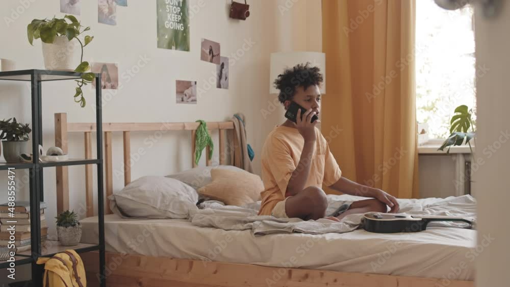 Stab shot of Biracial teenage boy talking with friend on mobile phone sitting on bed in his room Stab shot of Biracial teenage boy talking with friend on mobile phone sitting on bed in his room