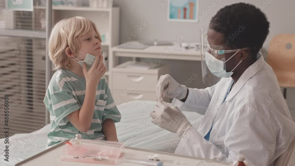 Medium slowmo shot of little Caucasian boy getting oral vaccination in doctors office given by young African-American male doctor in face shield and mask Medium slowmo shot of little Caucasian boy getting oral vaccination in doctors office given by young African-American male doctor in face shield and mask