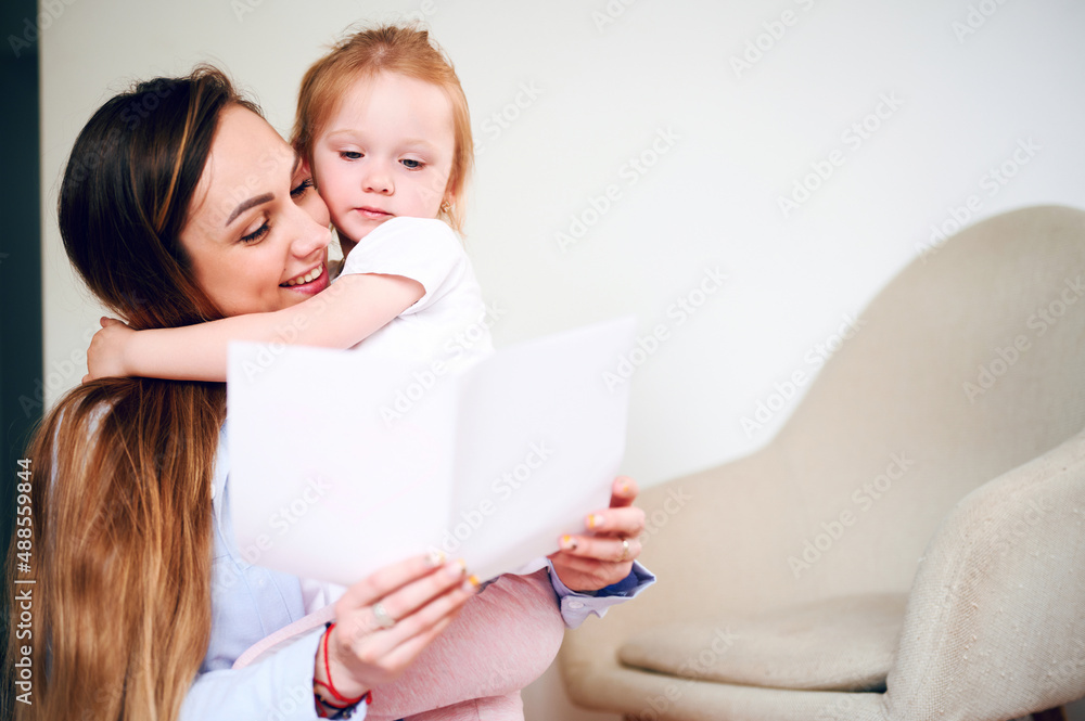Happy Mother's Day. Happy smiling mom with her little daughter reading holiday card while sitting on the floor Happy Mother's Day. Happy smiling mom with her little daughter reading holiday card while sitting on the floor
