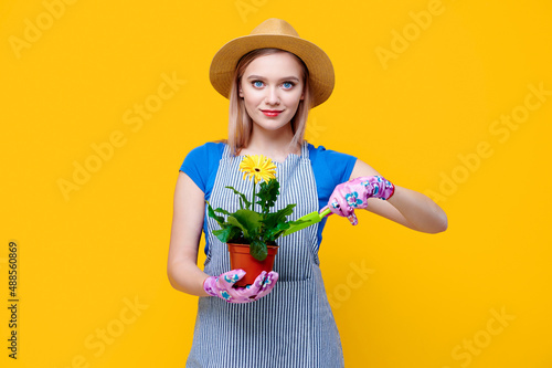 Young smiling caucasian woman holding flower gerbera garden gardener holding gerbera flower in her hands looks after it with spatula and rake on yellow background