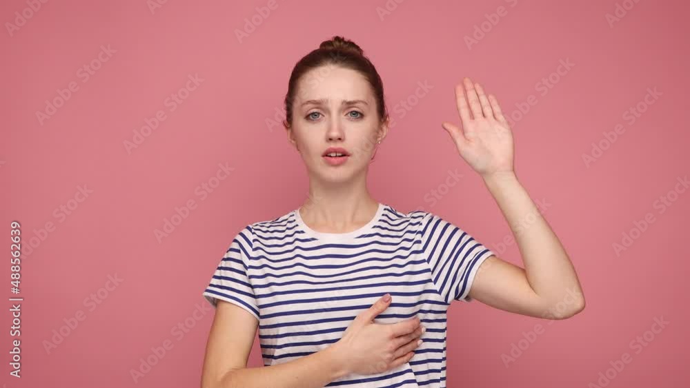 Serious conscious woman with hair bun holding one hand on chest and raising another swearing, trust and honest, patriotism, wearing striped T-shirt. Indoor studio shot isolated on pink background. Serious conscious woman with hair bun holding one hand on chest and raising another swearing, trust and honest, patriotism, wearing striped T-shirt. Indoor studio shot isolated on pink background.