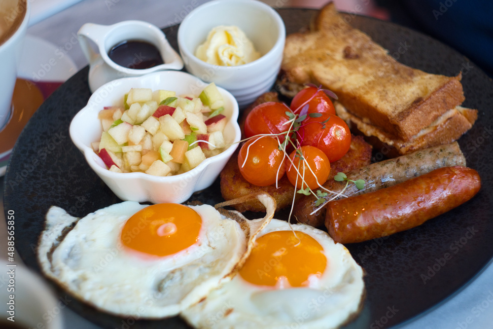 Full plate of traditonal English breakfast - fried eggs, toasted bread, grilled cherry tomato, sausage, beans, fruit salad with marple syrup Full plate of traditonal English breakfast - fried eggs, toasted bread, grilled cherry tomato, sausage, beans, fruit salad with marple syrup