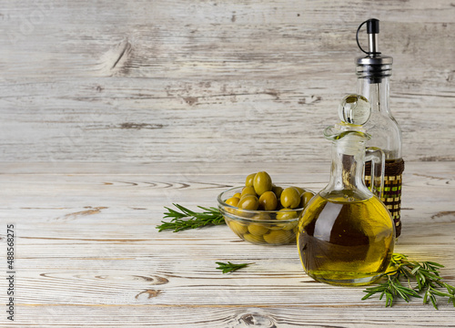 Fototapeta Naklejka Na Ścianę i Meble -  A jug and a bottle of olive oil on a light wooden table. Olives in a stalking vase and sprigs of rosemary in the background. Space for text