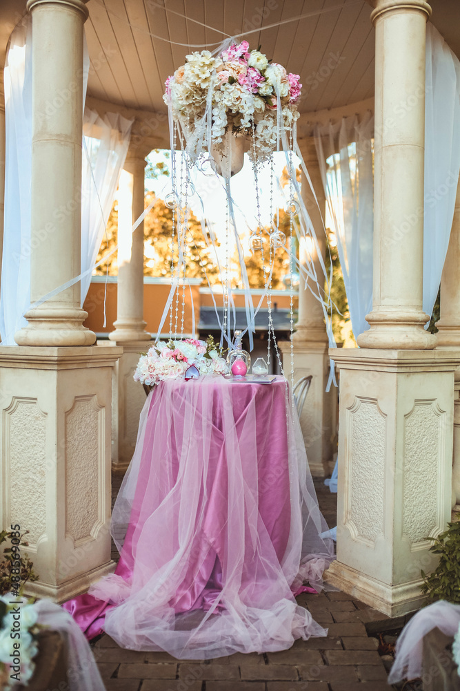 a flower arrangement of white and pink flowers, white ribbons and transparent beads under a cream-colored arch dome is fixed above a table covered with a pink cloth a flower arrangement of white and pink flowers, white ribbons and transparent beads under a cream-colored arch dome is fixed above a table covered with a pink cloth