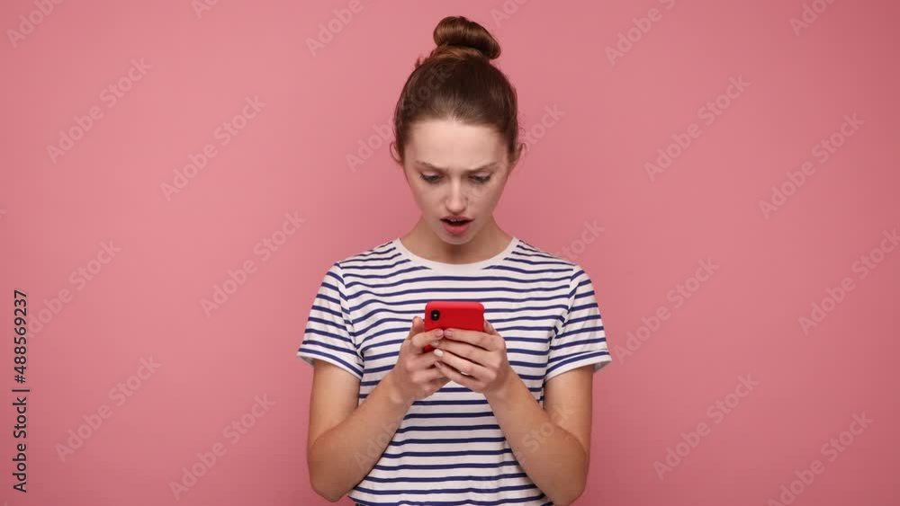 Shocked astonished woman with hair bun reading post on social network using cell phone, chatting, looking surprised, wearing striped T-shirt. Indoor studio shot isolated on pink background. Shocked astonished woman with hair bun reading post on social network using cell phone, chatting, looking surprised, wearing striped T-shirt. Indoor studio shot isolated on pink background.