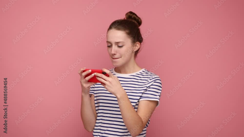 Happy excited woman using mobile phone and smiling, playing video game on cellphone, expressing excitement, wearing striped T-shirt. Indoor studio shot isolated on pink background. Happy excited woman using mobile phone and smiling, playing video game on cellphone, expressing excitement, wearing striped T-shirt. Indoor studio shot isolated on pink background.